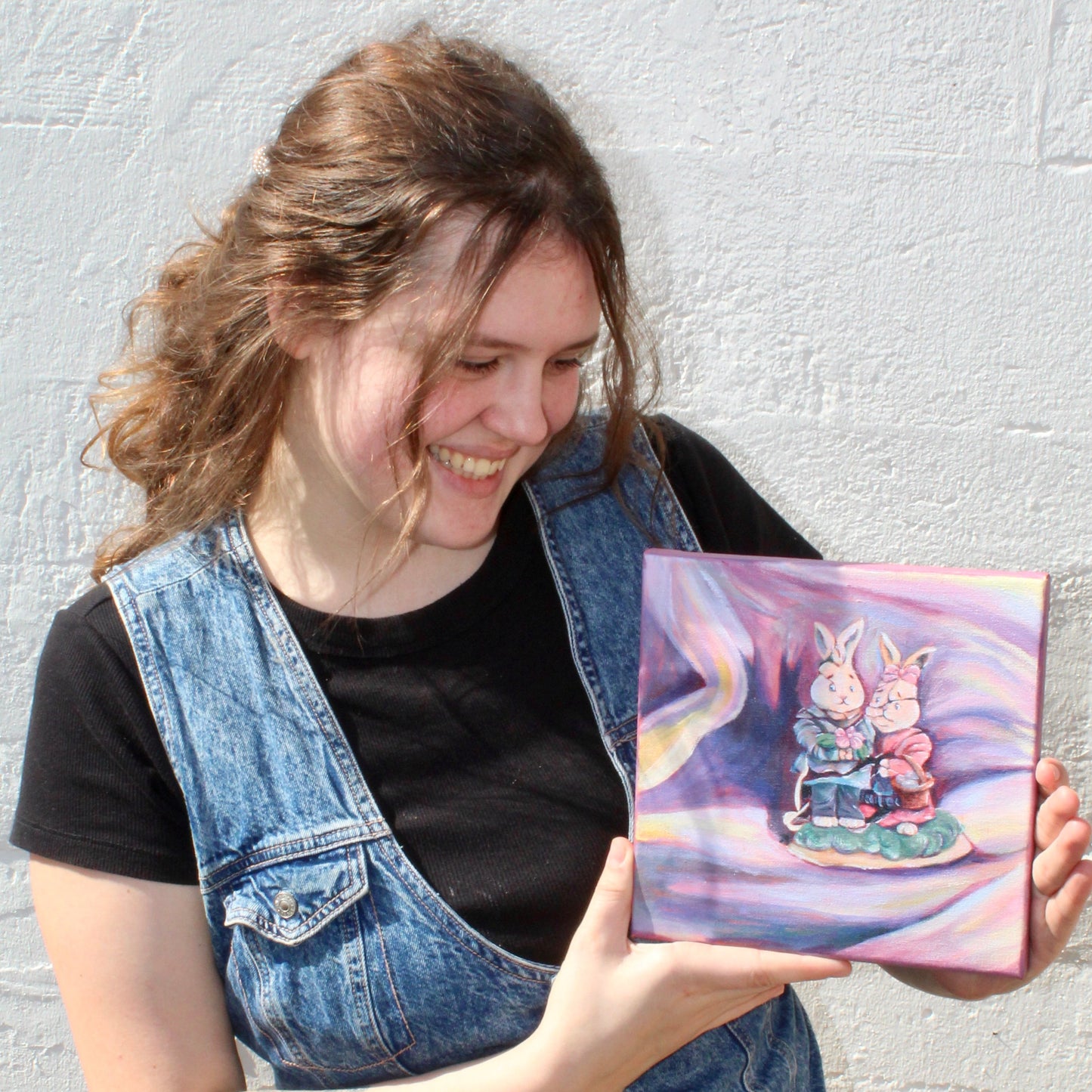 Resident artist, Bailey Crabtree, holding her painting of a broken rabbit figurine, titled "Broken Together/ Falling Apart." against a white wall